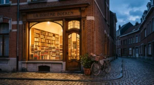 Étal en bois d'une devanture de librairie indépendante belge en brique rouge, avec piles de livres aux couvertures neutres sous une lumière douce de fin de journée
